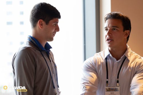 Two men in business casual discussing in a bright office by a window; one wears a lanyard and name badge reading Nico.