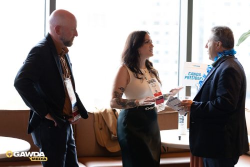 Three professionals in a lounge area with conference badges, conversing by a window; a GAWDA sign is visible in the background.
