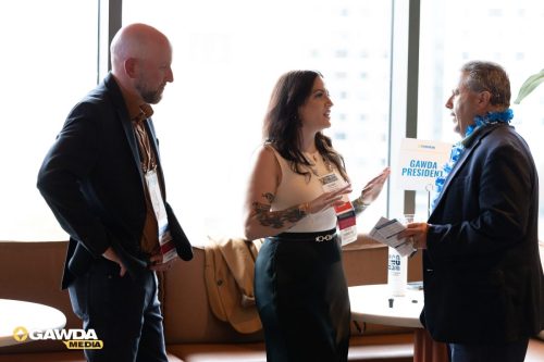 Three professionals in business attire converse near a 'GAWDA President' sign at a conference.