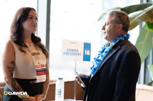 Woman with tattoos and a name badge converses with a man in a suit wearing a blue lei at a conference, GAWDA President sign in the background.