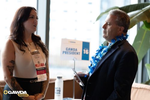 Two people talking at a conference: a tattooed woman with a name badge and a man in a suit wearing a blue lei, with a 'GAWDA PRESIDENT' sign in the background.