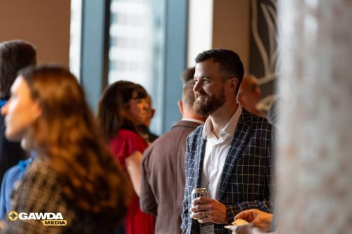 Man with beard in a checked blazer holding a can, chatting at a crowded indoor event, logo bottom left reads GAWDA MEDIA.