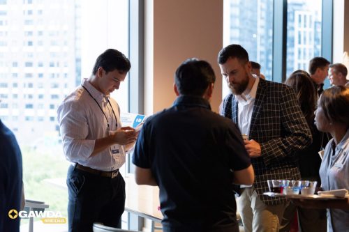 Group of professionals networking by a windowed wall at a daytime office event, with badges and drinks in hand.,