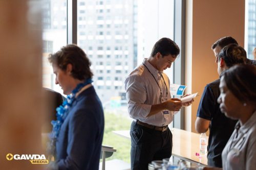 Man in a light-striped shirt with a lanyard reads a paper at a networking event by a large window overlooking city buildings. (GAWDA MEDIA logo visible bottom left)