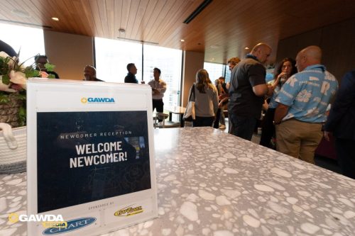Reception area with a sign reading 'Newcomer Reception — Welcome Newcomer!' at a GAWDA event, foreground shows a speckled table and floral arrangement with people chatting in the background.