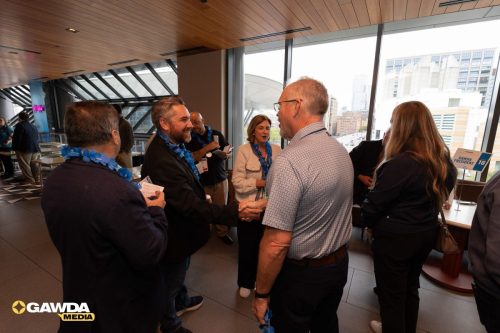 Group of professionals wearing blue leis shake hands and chat at a networking event in a glass-walled atrium; GAWDA Media logo in corner.