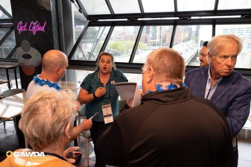 A woman in a teal shirt speaks to a small group of adults wearing blue leis and conference lanyards in a bright venue with city buildings visible through large windows.