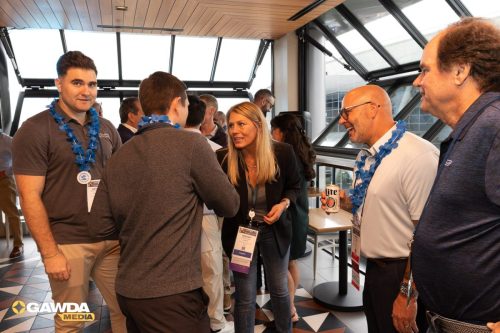 Group of professionals networking in a sunlit atrium, wearing blue lei garlands and name badges.