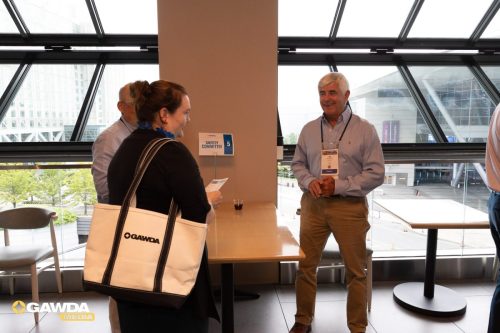 Woman with a white tote bag talking to a man in a light blue shirt at a 'Safety Committee' sign by a windowed wall, with a drink on the table nearby.
