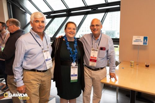Three conference attendees pose together by large windows, wearing lanyards and badges for the event.