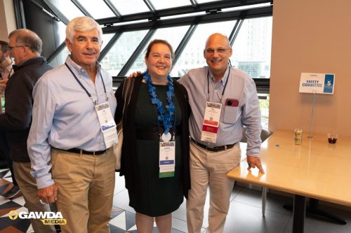 Three smiling adults wearing conference badges pose together at a daylight conference venue by a table with a 'Safety Committee' sign in the background.