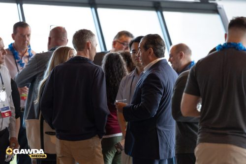 Group of professionals networking in a bright room, several wearing blue leis, as a man in a navy suit talks with another attendee with a drink in hand momentarily visible in the foreground, near a large windowed wall.