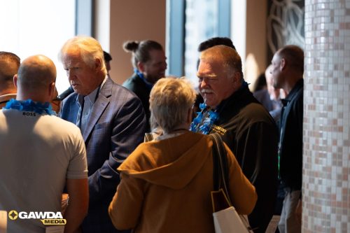 Group of people chatting at an indoor event, wearing blue flower leis and name badges; GAWDA Media logo visible in the corner.