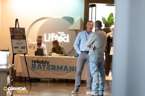 Registration desk at a conference booth with two staffers organizing papers as attendees chat nearby, sponsor signage visible.