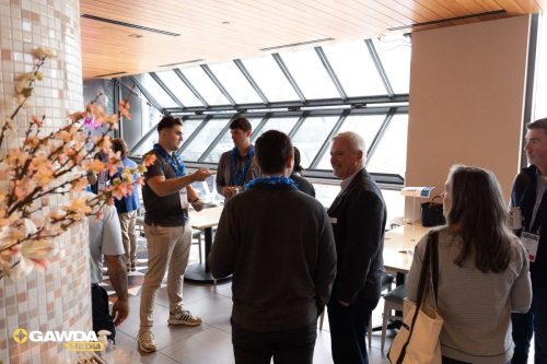 Group of professionals wearing blue leis chat and mingle near a large windowed wall at a conference reception.