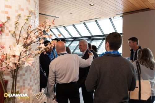 Group of professionals networking in a bright atrium with a mosaic column and pink blossoms; a man in a white shirt greets others nearby.