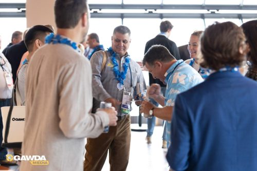 Group of professionals at a bright indoor event, wearing blue leis and name badges, chatting and holding drinks near a booth area covered with natural light.