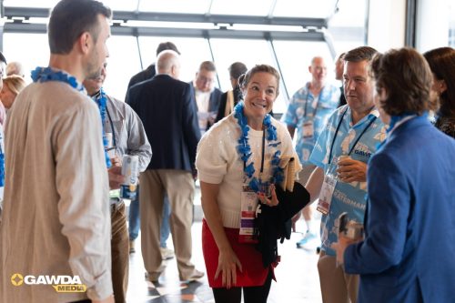 Group of conference attendees chatting in a bright venue; woman in white sweater and red skirt wears a blue lei and badge, smiling at the camera.