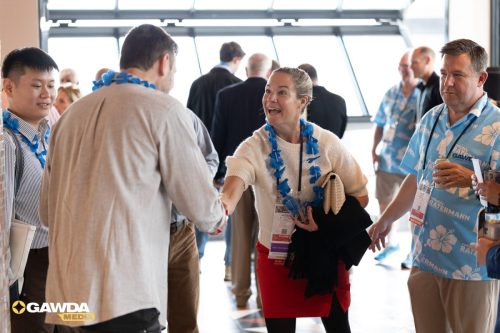 Woman in cream sweater greeting a man with a handshake at a networking event, wearing blue leis and a name badge; others chat in background near large windows.