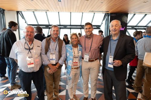 Five professionals posing for a group photo at a conference in a glass-walled atrium, all wearing name badges and casual business attire.