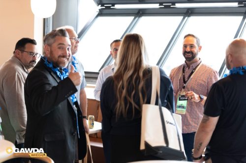 Group of professionals networking and chatting in a bright room with large windows; a man in a suit gives a thumbs up while others listen.