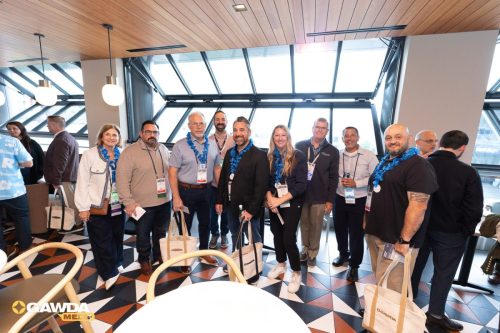 Group of smiling professionals with blue leis posing for a group photo at a conference, holding tote bags and badges in a modern venue.