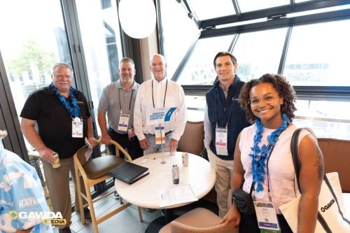 Five people with conference badges and blue leis pose around a round table by large windows, smiling at the camera; drinks and papers are on the table.