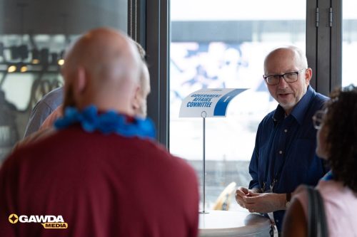 Man with glasses in a blue shirt speaks to attendees at a counter beside a 'Government Affairs Committee' sign.