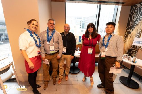 Five people wearing blue leis pose for a group photo at a conference lounge near large windows in a high-rise venue.