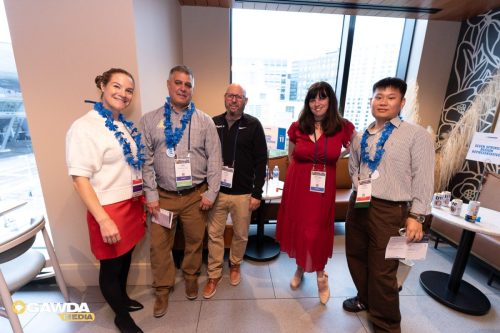Five attendees wearing blue leis and name badges pose for a group photo in a modern conference area with large windows.