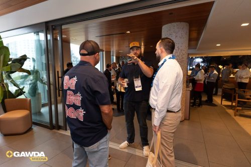 Three men chat in a hotel lobby during a conference; one in a navy polo with lanyard, another in a check shirt, background attendees nearby. GAWDA Media logo visible in corner.