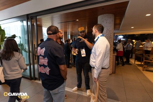 Networking event in a modern lounge with attendees chatting in small groups; a man in a navy shirt with large red lettering on the back stands with others near a doorway, badges visible around their necks.