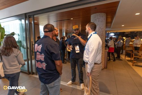 Group of men conversing in a modern lobby area near glass doors during a networking event; one man wears a navy shirt with large red 'Bat' text and a cap, others have lanyards.