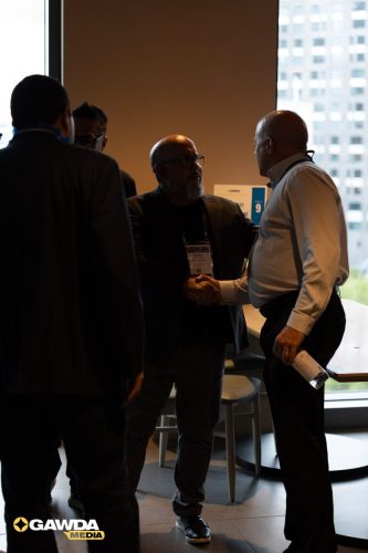Two men shake hands in a dim conference space by a window, with name badges and the GAWDA Media logo in the corner.
