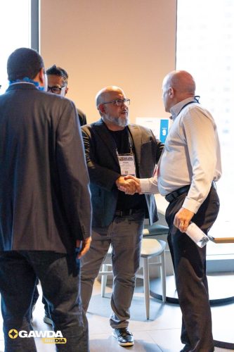 Two men shake hands in a conference lobby while others converse nearby; badges visible on the central man with glasses and beard, GAWDA MEDIA logo bottom-left