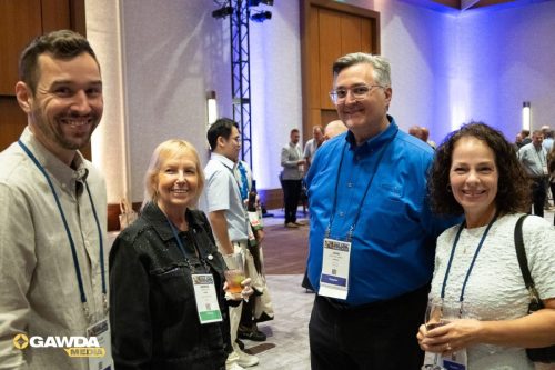 Four conference attendees pose for a group photo, wearing name badges, in a blue-lit event hall.