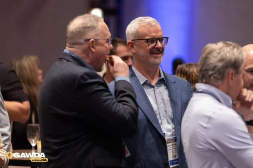 Smiling man in a light blue shirt and blue blazer speaks with a colleague at a networking event, with other attendees in the background and a visible name badge.
