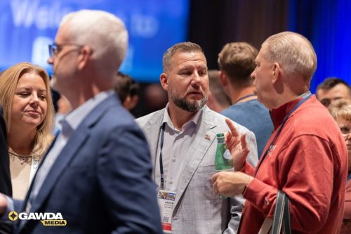 Professionals converse at a conference; man in a light gray blazer with a badge speaks with a man in a red shirt holding a bottle, background shows blue stage lighting and attendees.