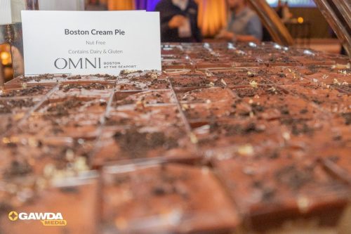 Tray of chocolate Boston Cream Pie bars on a display table, with a sign reading 'Boston Cream Pie' and allergen notes (Nut Free; Contains Dairy  Gluten).