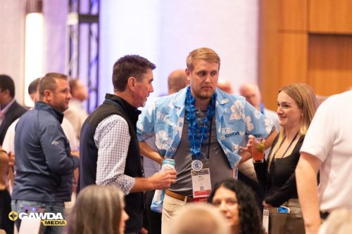 Group of conference attendees chatting at a networking event; a man in a blue Hawaiian shirt with a flower lei and badge stands with two others, drinks in hand.
