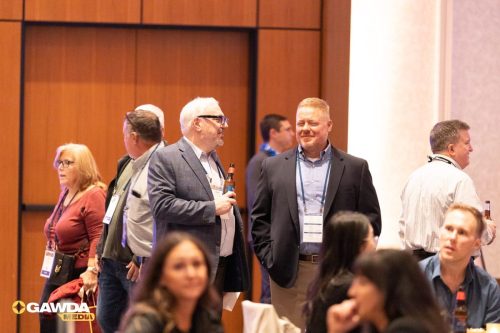 Professionals mingle at a conference, two men in the foreground chatting in suits with name badges and lanyards, GAWDA Media logo visible.