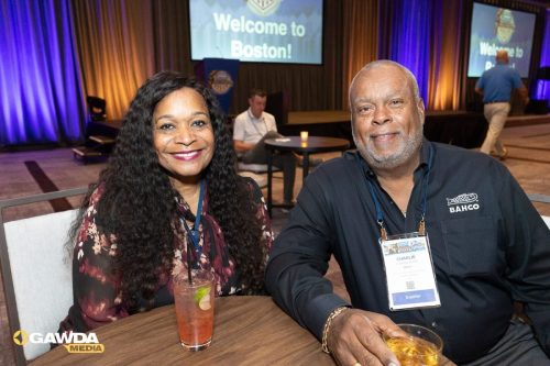 Two smiling attendees sit at a round table with drinks, wearing conference badges, in a venue with a 'Welcome to Boston!' screen in the background.