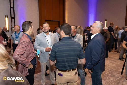 Professionals networking at a conference reception, talking in groups with drinks in hand and badges visible, GAWDA Media watermark bottom left.