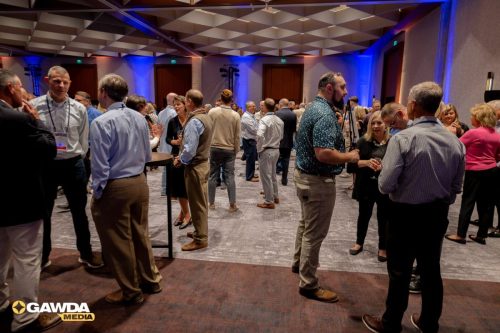 Crowd of professionals chatting at a conference reception in a modern venue with blue uplighting and name badges.