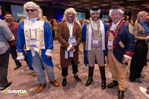 Five men in 18th‑century costumes with wigs and name badges posing at a conference.
