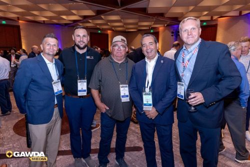 Five men in suits and casual wear pose for a photo at a conference, each with a name badge on a lanyard.
