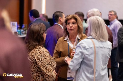 Middle-aged woman in a tan cardigan talks with another guest at a conference, holding a bottle; name badge visible at neck level.