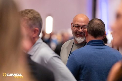 Two men chat at a crowded event; one with glasses and a gray beard faces a man in a blue shirt, with blurred attendees in the background.