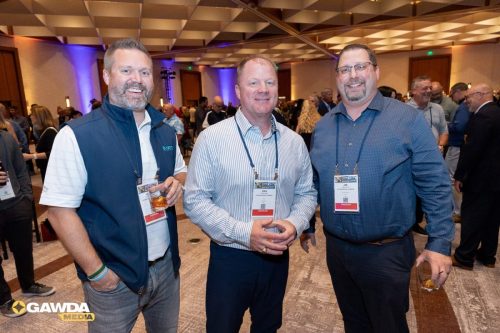 Three men with conference badges posing for a photo at a busy indoor networking event, drinks in hand and smiles visible, background attendees.