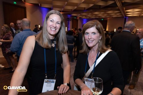 Two smiling women at a networking event with name badges standing at a high-top table, one holding a wine glass.